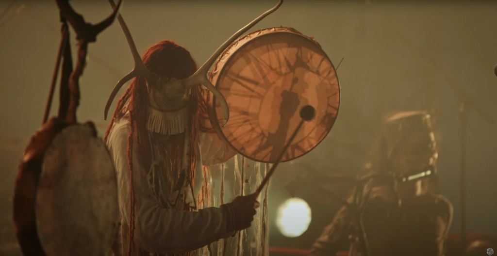 Video still from Heilung's performance. A drummer wearing a mask and horns beats a skin drum.