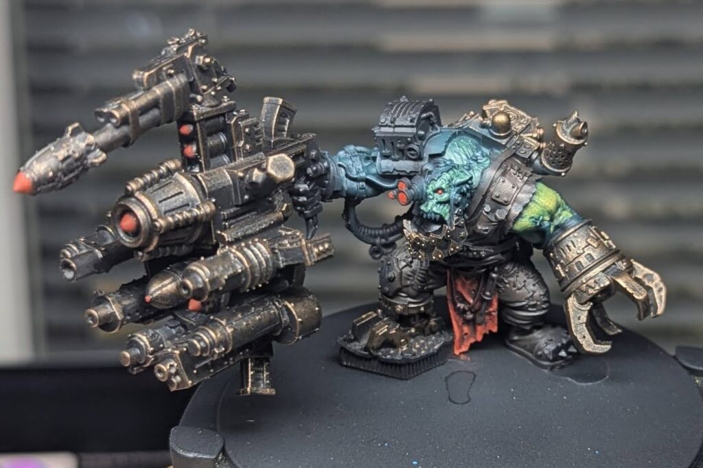 WIP photo of the MaxDakka ork painting. The skin has been airbrushed blue, and some green and yellow layers have been applied in parts. Red basecoats are present. Most of the model is dark brown over black. Metallic parts have been heavily drybrushed with a bronze colour.